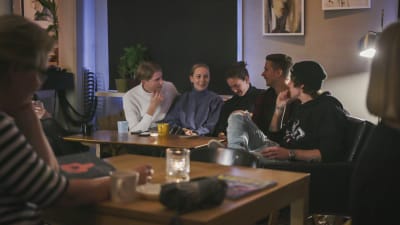 Fem unga personer sitter i en soffa på ett kafé och pratar.