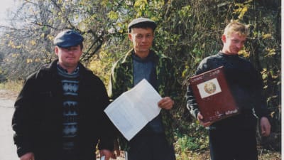 Tre män står utomhus och håller i papper tittar in i kameran. De här männen åkte runt i Vitryssland och hjälpte människor att rösta.