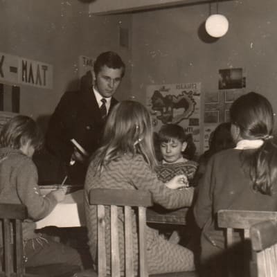 Nuori miesopettaja luokassa pulpeteissaan ahertavien pikkulasten keskellä, vanha valokuva.