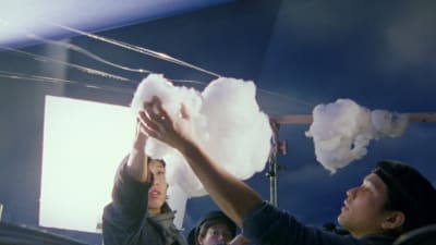 Kaksi miestä pitää kurottaen kiinni pumpulisesta pilvestä, joka on ripustettu pyykkinarua muistuttavaan laitokseen, taustalla sininen seinä ja suuri nelikulmainen kuvausvalo, kuva Hirokazu Kore-edan elokuvasta Elämän jälkeen.