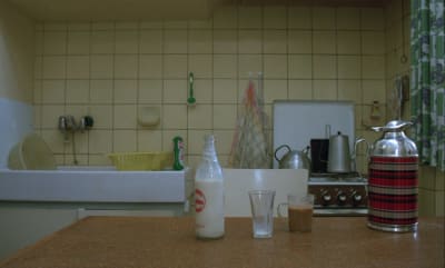 Maitopullo, lasi, kuppi ja termoskannu keittiön pöydällä, taustalla keittiön seinää, hella ja tiskipöytä.