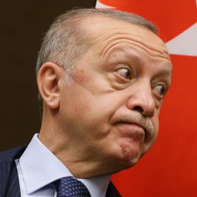Recep Tayyip Erdoğan tittar åt sidan och gör en grimas, han ser besviken ut – mungiporna pekar neråt. I bakgrunden syns Turkiets flagga.