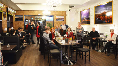 Flera personer i en restaurang runt ett långt bord. De flesta tittar mot någonting lite ovanför kameran.