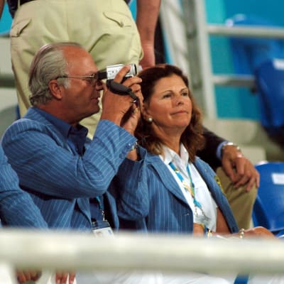 Kuningas kuvaa kameralla urheilukatsomossa. Kuninkaan toisella puolla istuu Carl Philip ja toisella puolella istuu Silvia.