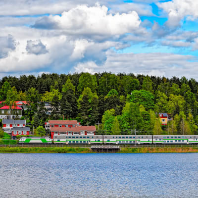 Kuva suomalaisessa maisemassa kulkevasta junasta. Metsää takana, vettä edessä. 