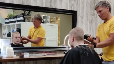 En ung man sitter i en frisörstol. Hans hår rakas bort av en frisör. På golvet en del hår.