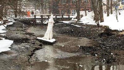 En å som är nästa utan vatten. Det snöar. En vit skulptur i vattnet. I bakgrunden en bro där två personer står. 