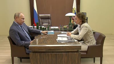 Rysslands president Vladimir Putin och barnombudsman Anna Kuznetsova sitter mittemot varandra vid ett skrivbord och ser allvarliga ut. 