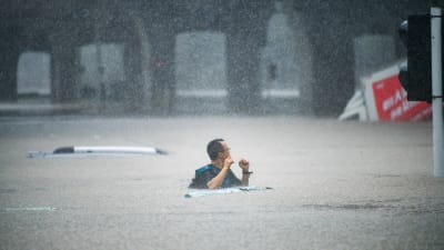En man går på en översvämmad gata där vattnet når honom ända till bröstet. I bakgrunden en lastbil som fastnat i vattenmassorna.