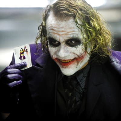 Heath Ledgerin esittämä Jokeri virnistää ja näyttää pelikorttia, jossa on jokerin kuva.