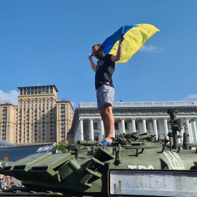 En man står på en förstörd rysk stridsvagn. Han håller upp den ukrainska flaggan.