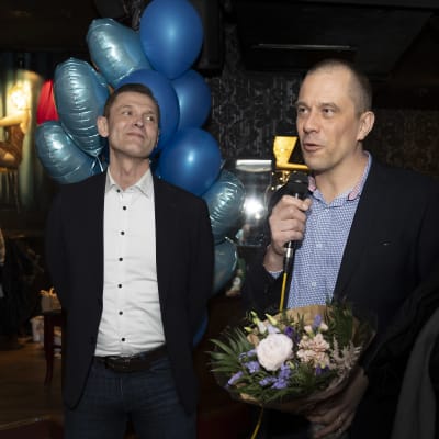 Sinuhe Wallinheimo kuuntelee hymyillen, kun Ville Väyrynen puhuu kukkakimppu kädessään Kokoomuksen vaalivalvojaisissa.