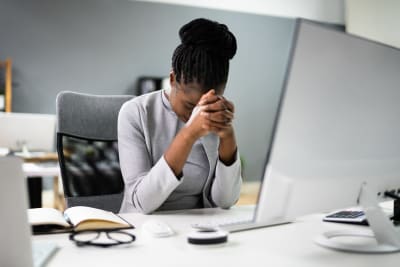 En kvinna sitter med händerna knäppta framför ansiktet och tittar ner i ett arbetsbord. På bordet finns en dator.