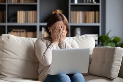 Kvinna sitter i en soffa med datorn i famnen, hon gömmer ansiktet i händerna av skam