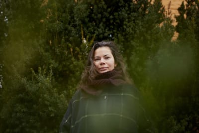 Liza Alexandrova-Zorina står bland gröna barrträd. Hon har en rutrandig jacka och en halsduk på sig.