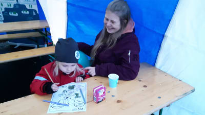 En kvinna och en liten flicka sitter vid ett bord inne i ett tält. Flickan färglägger en målarbild.