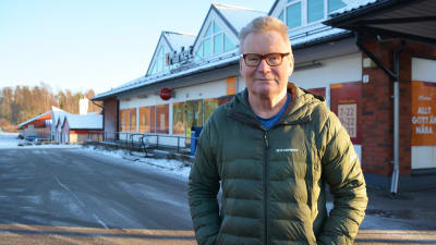 En man i grön dunjacka står utanför en matbutik i ett snöigt landskap. Han har händerna i fickorna och tittar in i kameran.