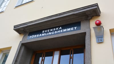 Närbild på en gul byggnad med flera fönster. Ovanför dörren i trä står det Svenska församlingshemmet med versaler.