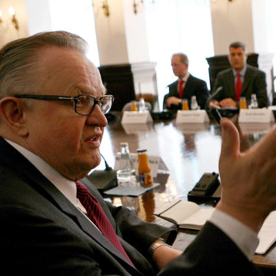 Martti Ahtisaari sitter vid ett stort förhandlingsbord där alla män i kostym har en namnskylt framför sig. Han pratar med någon sin inte syns i bild och gestikulerar.