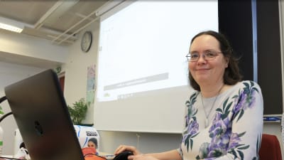 En kvinna sitter vid en dator med en stor belyst skärm på väggen bakom henne. Hon småler in i kameran.