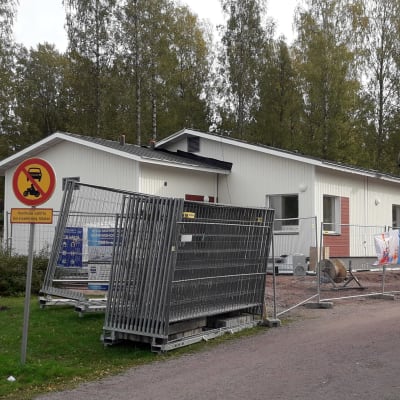 Vita byggnader med tillfälliga stängsel rumtomkring. En skylt som visar att det är förbjudet att klöra in på området. Grusväg mellan byggnaderna och träd i bakgrunden.