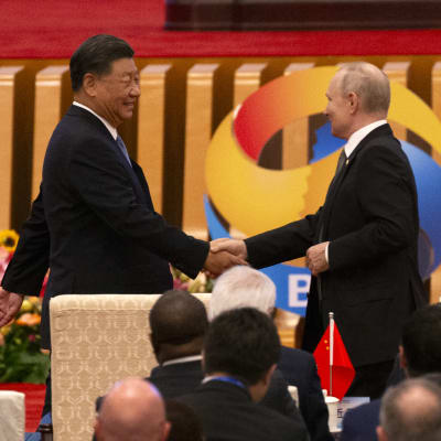Kiinan presidentti Xi Jinping ja Venäjän presidentti Vladimir Putin kättelevät kokoussalissa, etualalla muita osallistujia.