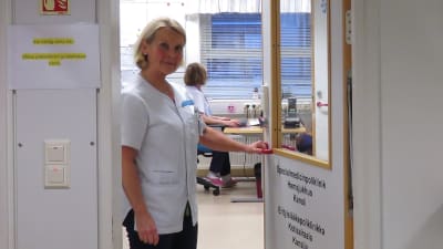 En kvinna i sköterskekläder står vid en innerdörr till ett kansli. Det står bland annat hemsjukhus på dörren.