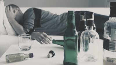 Man ligger i en soffan med handen för ansiktet och flera tomma spritflaskor på bordet.