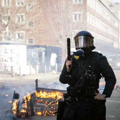 Polis med hjälm och gasmask håller ett vapen i handen och på gatan brinner något.