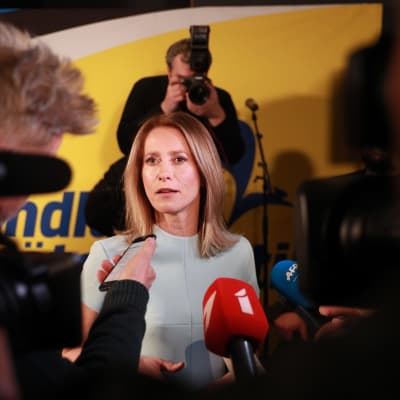 Pitkätukkainen nainen lyhythihaisessa mekossa antaa haastatteluja kameroiden ja mikrofonien ympäröimänä.