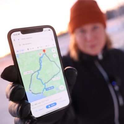 Toimittaja näyttää kameralle puhelinta, jossa näkyy Google Mapsin reittivaihtoehdot välillä Oulu–Rovaniemi.