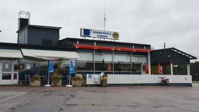 Ett svart hus med ett vitt lågt staket utanför. Framför huset syns asfalt och bakom huset grå himmel. På taket finns en skylt där det står Koskenkylä Forsby med en bild av en kaffekopp.