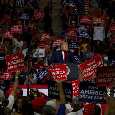 President Donald Trump står vid ett podium i ett folkhav under ett kampanjmöte. Folk håller skyltar i händerna där det står Make America Great Again.