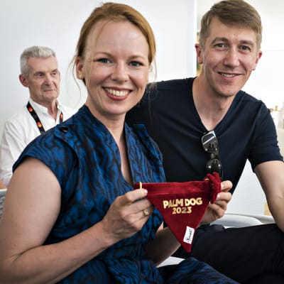 En glad Alma Pöysti håller upp en röd duk med texten "Palm Dog 2023" och bredvid henne ler Jussi Vatanen.