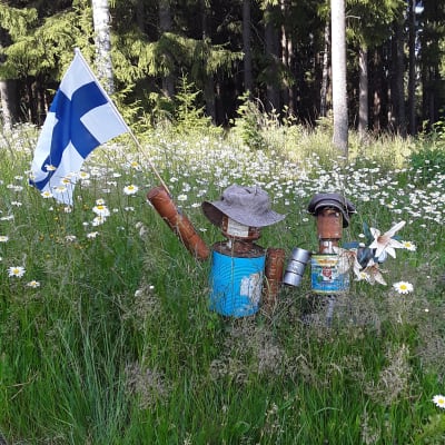 Kaksi peltiämpäreistä ja tölkeistä tehtyä hahmoa heinikossa. Toinen hahmoista tervehtii ohikulkijaa Suomen lippu kädessä.