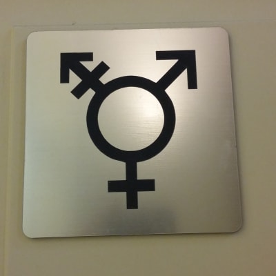 En toalettdörr med en skylt som visar att både män, kvinnor och transpersoner är välkomna att använda toaletten.