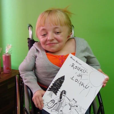 Monitaituri Noora Västinen istuu pyörätuolissaan. Hänellä on kädessään Roisku ja Loisku -lastenkirja, jonka hän on itse kirjoittanut ja kuvittanut. 