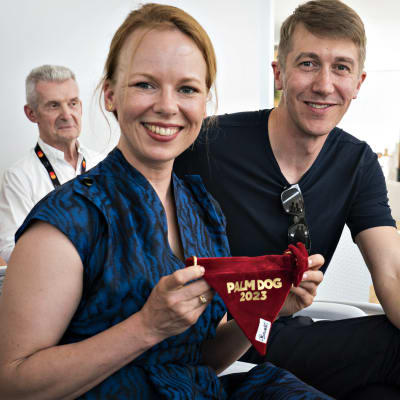 En glad Alma Pöysti håller upp en röd duk med texten "Palm Dog 2023" och bredvid henne ler Jussi Vatanen.