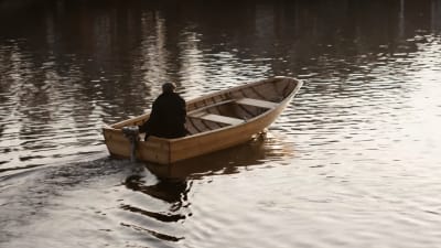 Man sitter i en båt med aktersnurra och åker bortåt från fotografen