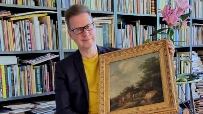 Författaren Peter Mickwitz sittandes framför en bokhylla med en gammal oljemålning i famnen