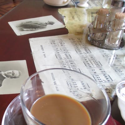 Keskeneräinen kahvikuppi kahvilan pöydällä, jolla nuotteja ja valokuvia.