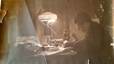 En hundra år gammal bild av en man som sitter och skriver vid sitt arbetsbord.