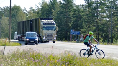En cyklist cyklar på en riksväg. Bakom cyklisten skymtar en stor långtradare.
