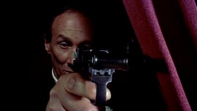 Mies (näyttelijä Reggie Nalder) tähtää pistoolilla punaisen verhon takaa melkein kohti kameraa. Kuva elokuvasta Mies joka tiesi liikaa.