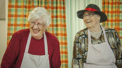 Två äldre damer klädda i förkläden, den ena i svart hatt, skrattar.