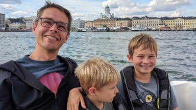 Olof och hans två barn sitter i en båt med Helsingfors i bakgrunden.