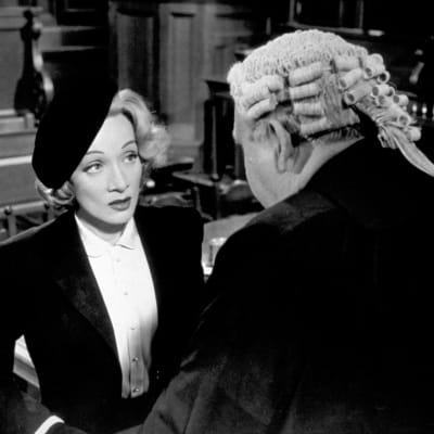 Baskeripäinen Christine Vole (Marlene Dietrich) peruukkipäisen asianajajan (Charles Laughton, selin) puhutettavana oikeussalissa. Kuva elokuvasta Todistaja (Witness for the Prosecution)