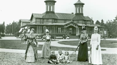 Fyra kvinnor i långa klänningar och fina hattar framför en utsmyckad träbyggnad. Mellan dem sitter tre barn i gräset. Kvinnorna håller alla i tennisracketar. Bilden är svartvit och från 1910.