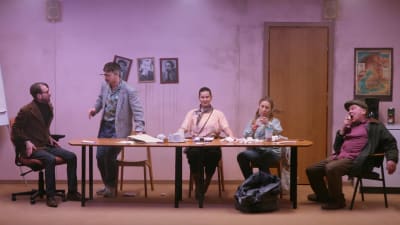 Fem skådespelare sitter bakom ett långt mötesbord i ett sjabbigt kontor. 