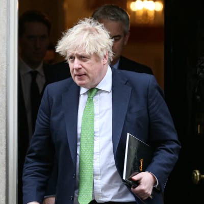 En rufsig blond man i grön slips med en mapp i handen går ut genom en svart dörr.